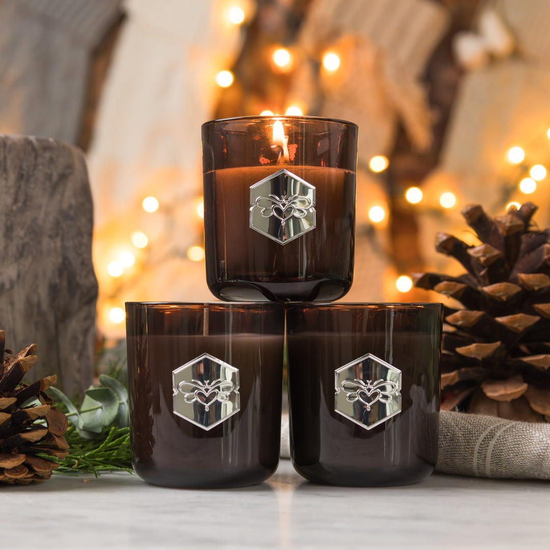 Three dark glass candles with a logo on a wooden surface with blurred lights and pinecones in the background