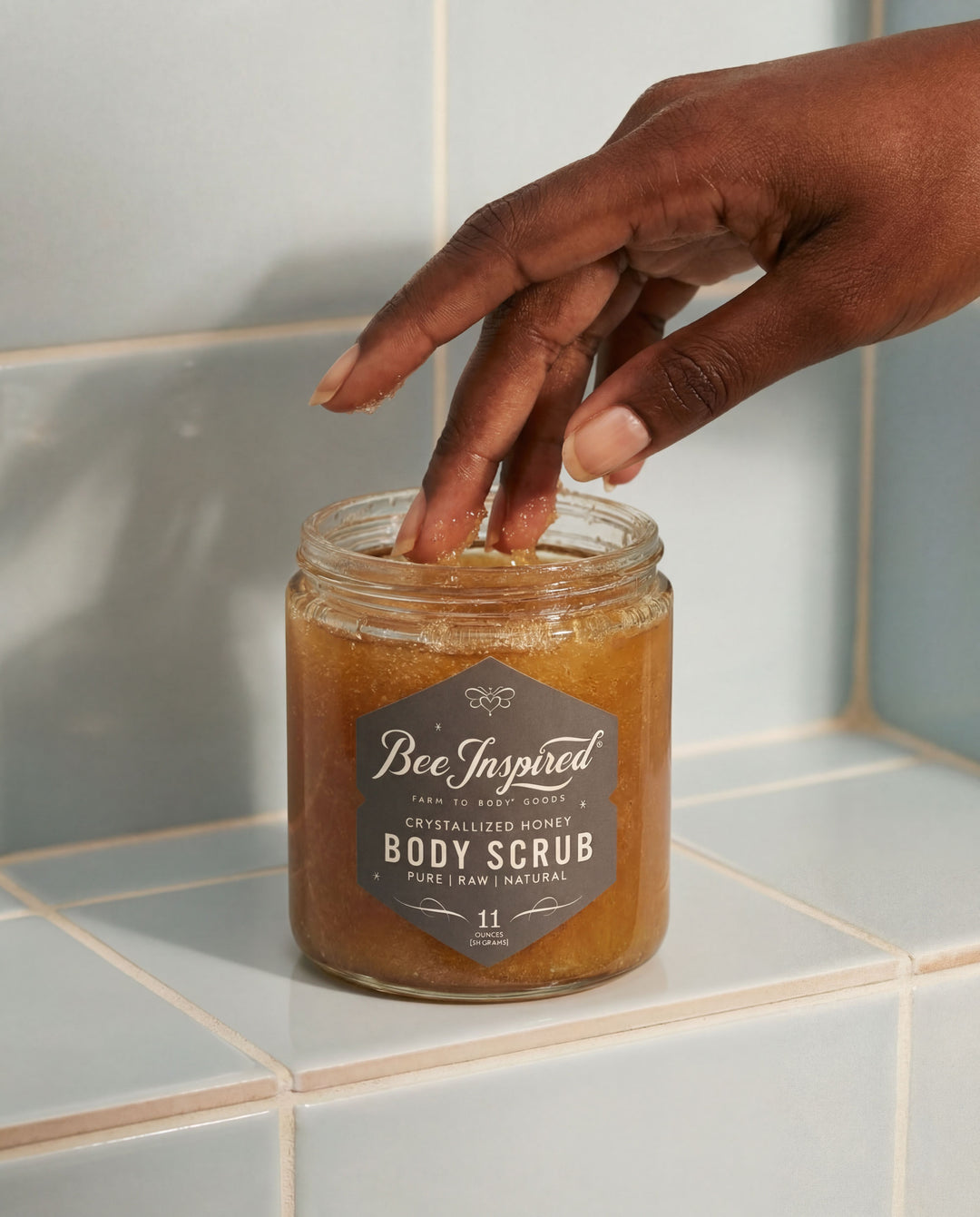 Jar of Bee Inspired body scrub on a tiled bathroom shelf with a hand reaching towards it.