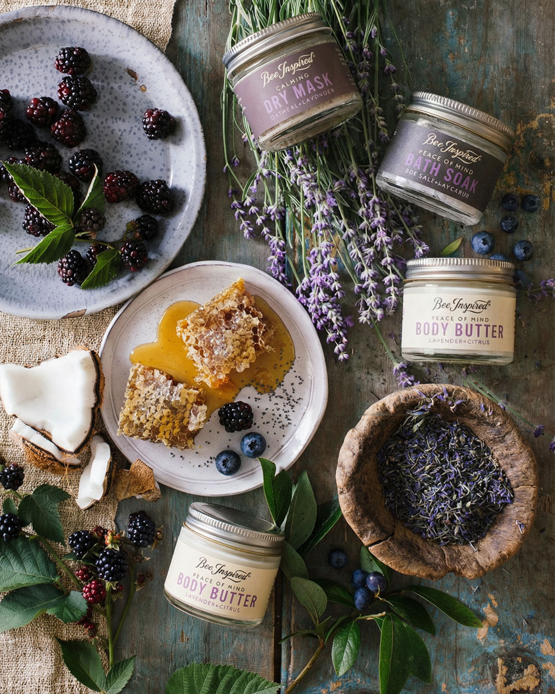 Assorted natural beauty products including jars of body butter, a bowl of lavender, and a plate with honey and blackberries on a rustic wooden surface.
