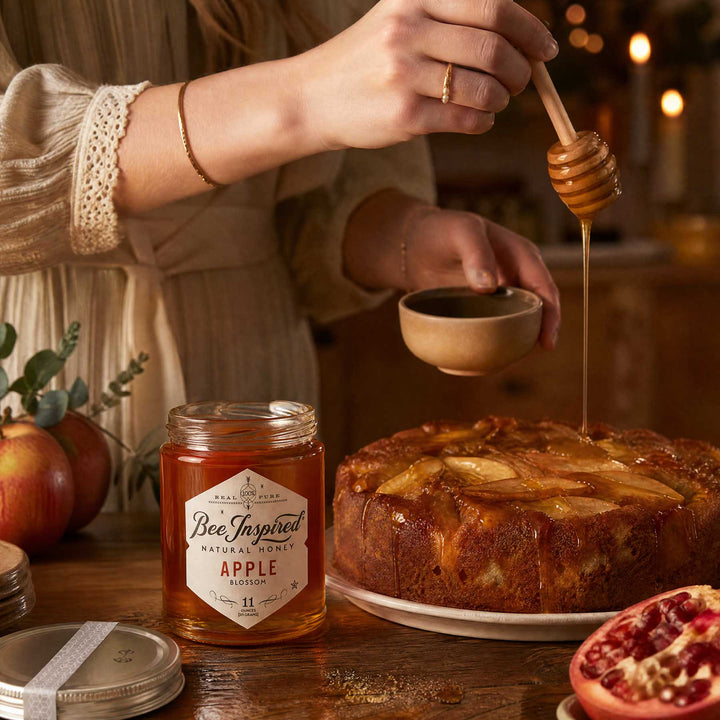 A person in a cream dress drizzles honey from a wooden dipper over an apple pie on a wooden table, with a jar of Bee Inspired Natural Honey Apple Blossom in the foreground.