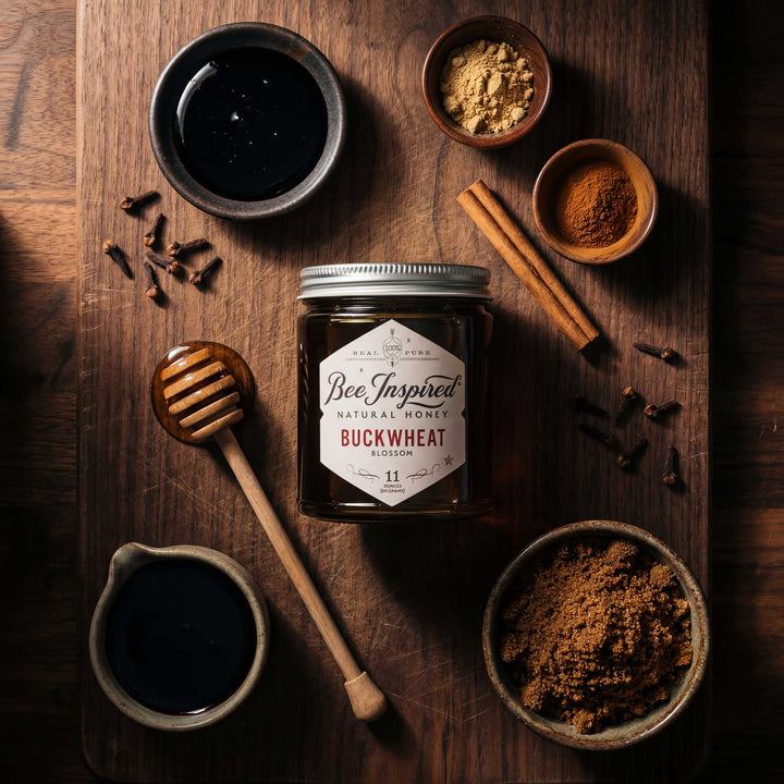 Jar of Bee Inspired Buckwheat honey on a wooden surface with spices and a honey dipper.