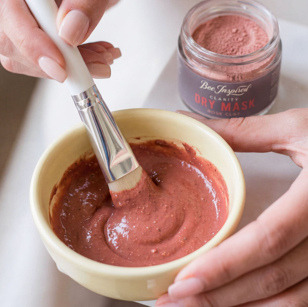 Person mixing a pink clay mask in a bowl with a brush, next to a jar of the same mask.