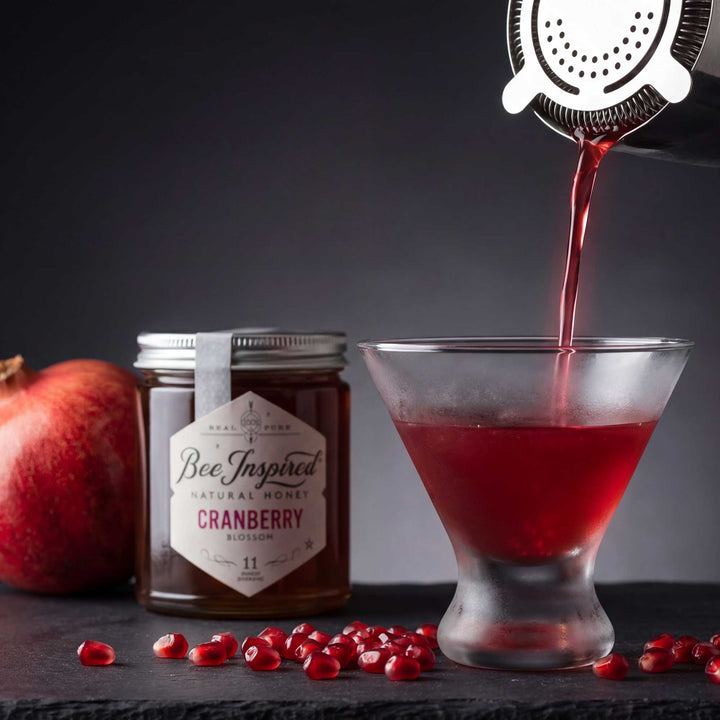 Cranberry juice being poured into a glass with a jar of cranberry honey and pomegranate fruit on a dark surface.