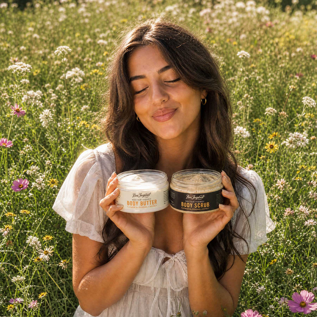 A woman in a white blouse stands in a sunlit field of wildflowers, holding two jars of The Body of Thought body care products: a white jar labeled Body Butter on the left and a dark brown jar labeled Body Scrub on the right.