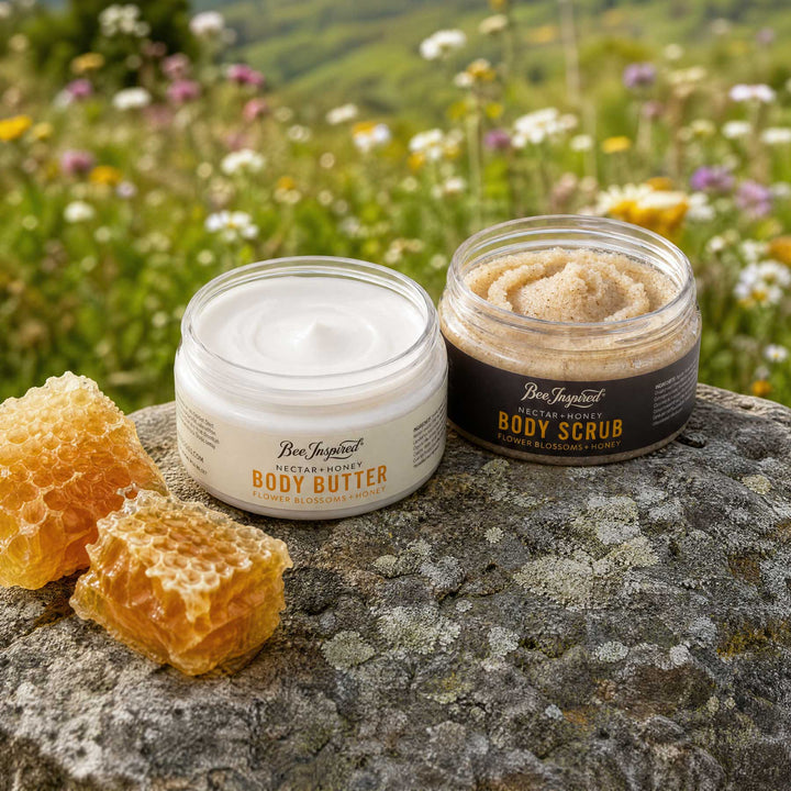 Two jars of Bee Inspirational body skincare products, Body Butter and Body Scrub, displayed on a stone with honeycomb in a blurred meadow background.
