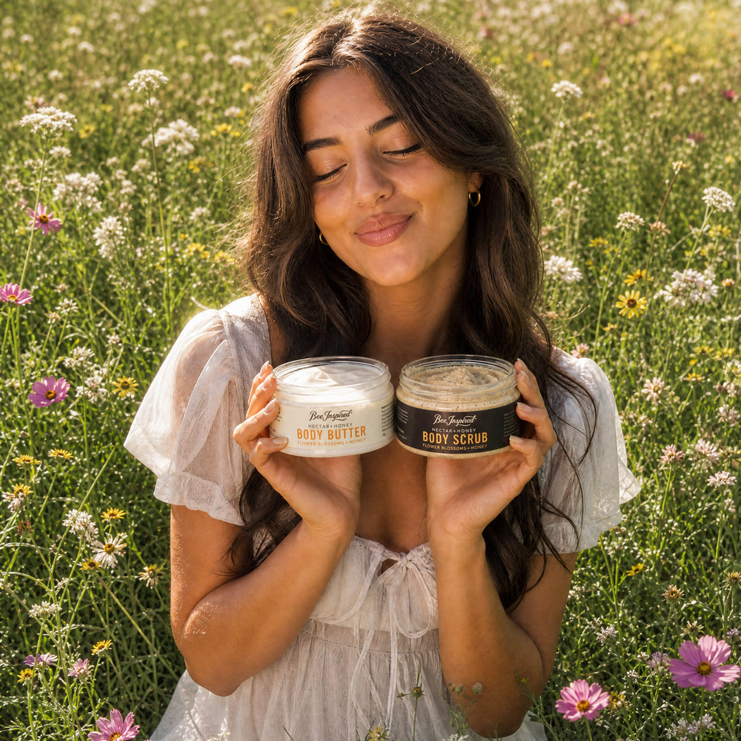 A woman in a white ruffled dress stands in a sunlit wildflower field, holding two jars of body care products: one labeled body butter and one labeled body scrub.