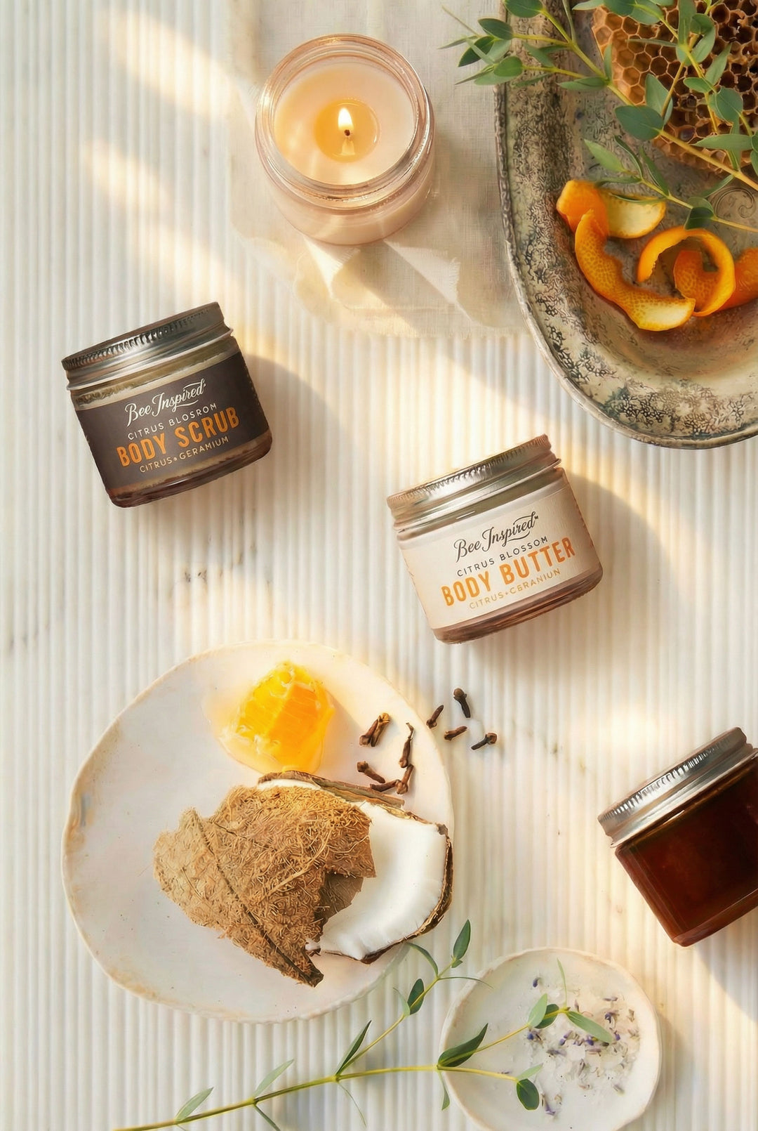 Body care products including body scrub, body butter, and a candle on a textured surface with decorative elements.