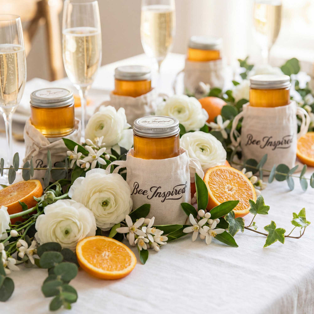 Jars of honey with 'Bee Inspired' labels on a table with flowers and oranges.