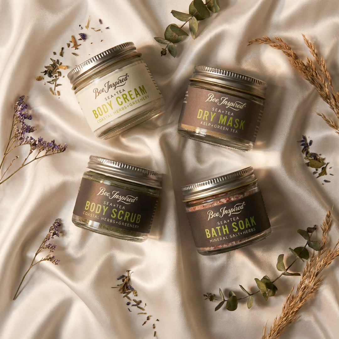 Four jars of Bee Inspired body care products on a textured fabric background with herbs and flowers.