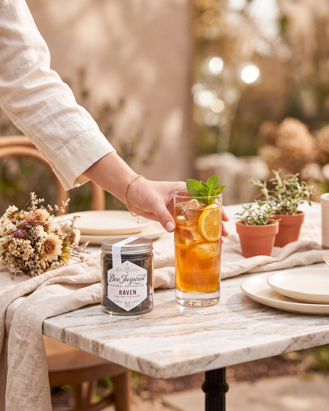 Person holding a glass of iced tea with a jar of Bee Inspired Raven Tea on a table outdoors.