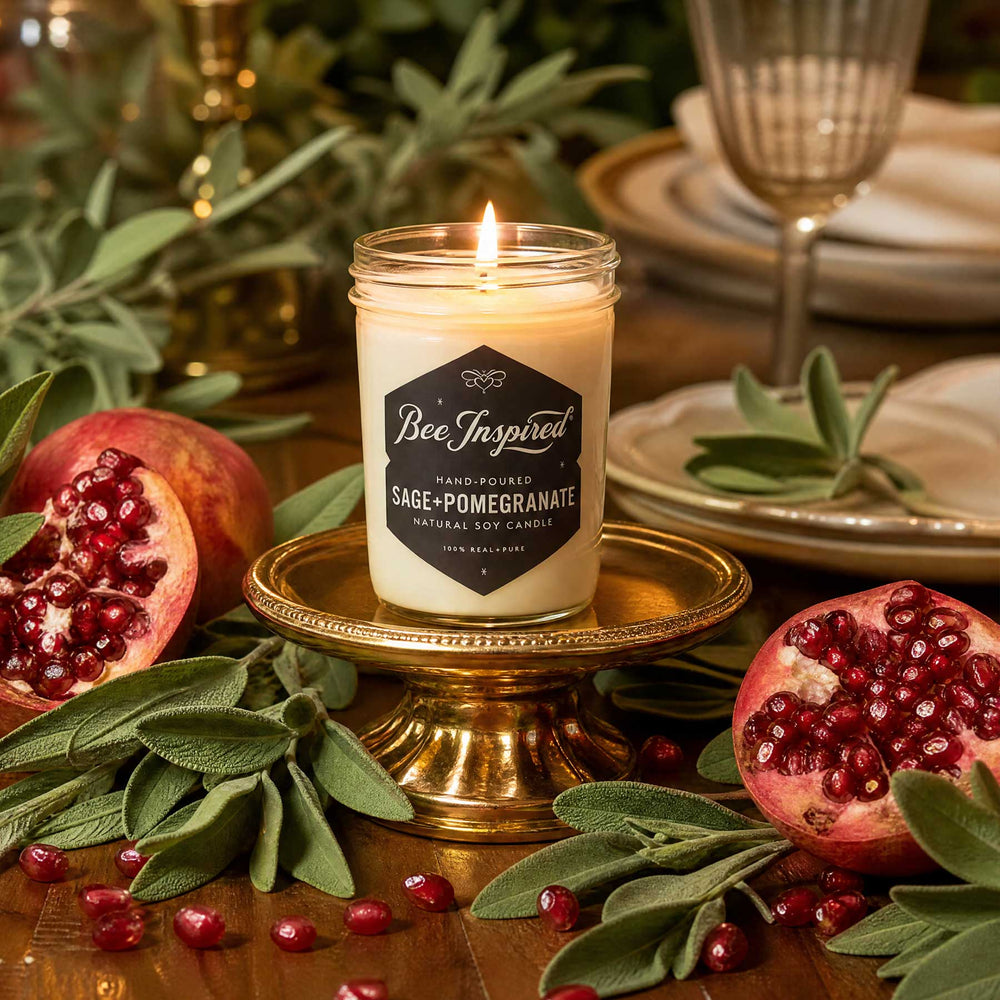 A lit candle in a clear glass jar sits on a gold pedestal, surrounded by pomegranates, sage leaves, and a warm, festive tabletop setting. The candle label reads 'Bee Inspired' and 'Sage-Pomegranate'.