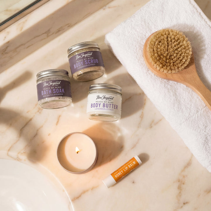 Bee Inspired skincare products including body scrub, body butter, and lip balm on a marble surface with a candle and brush.